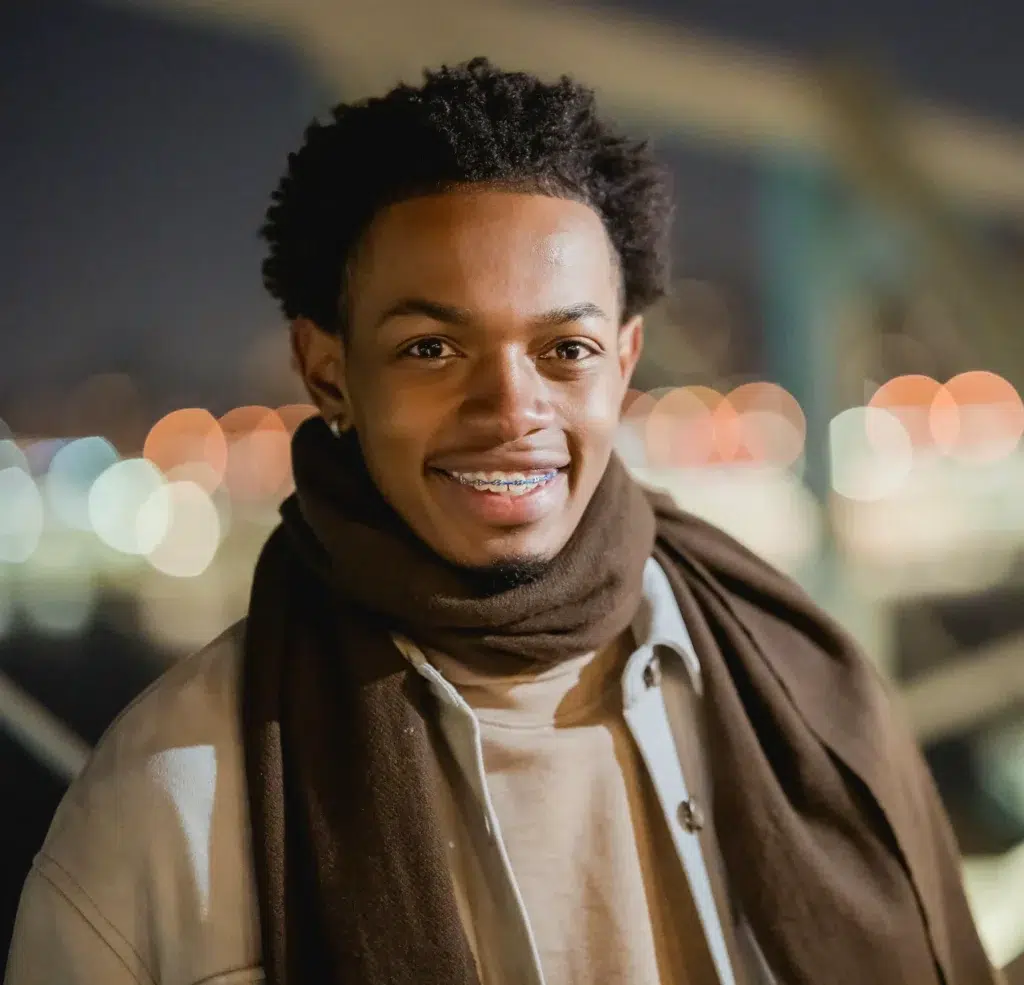 Smiling young man with braces wearing a scarf and jacket at night with blurred city lights in the background.